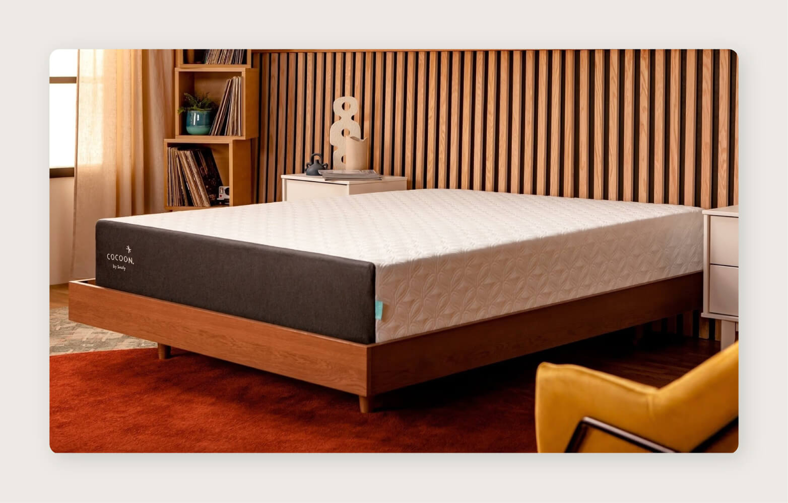 A Sealy Cocoon Chill Mattress on a platform frame in a retro bedroom with a wood slat wall, burnt orange rug, and yellow chair. 