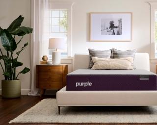 The Purple RestorePlus™ mattress on a cream upholstered bed frame between two wood nightstands in a bright bedroom.