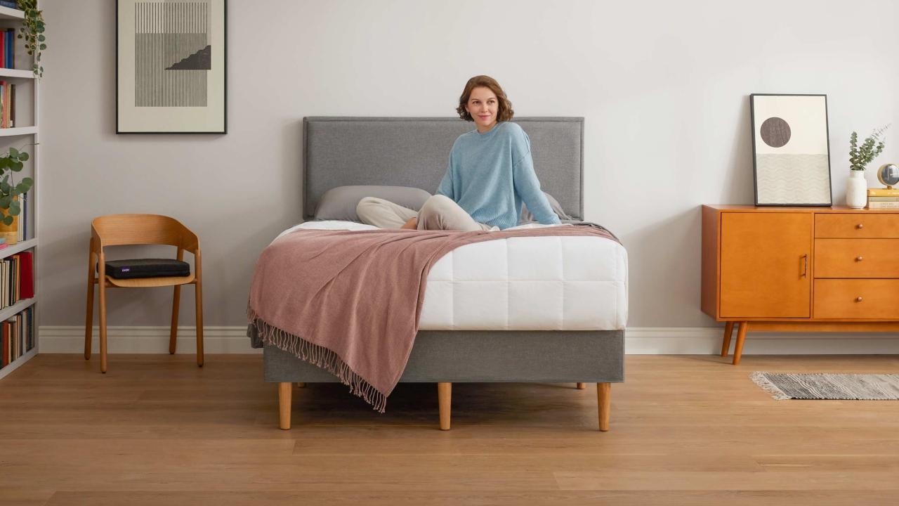 A woman sits upright on top of the covers of a bed on the Purple Bed Frame.