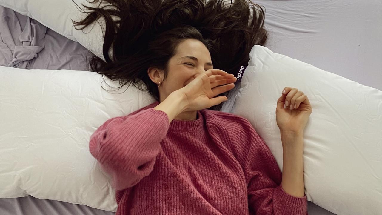 Woman lying on her back surrounded by three Purple pillows. 