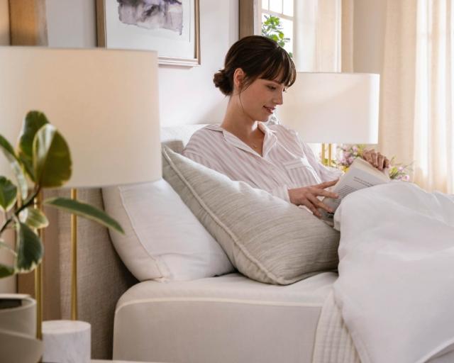 A woman reads a book in a bed with white sheets next to a green plant.