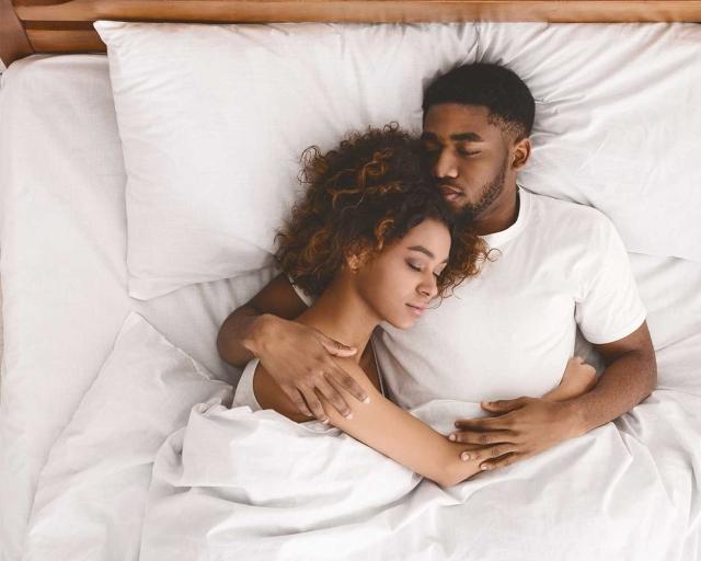 A couple cuddling together in a california king bed with white sheets and white pillows. 