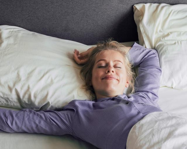 A woman wearing purple pajamas wakes up smiling in a bed with white sheets.