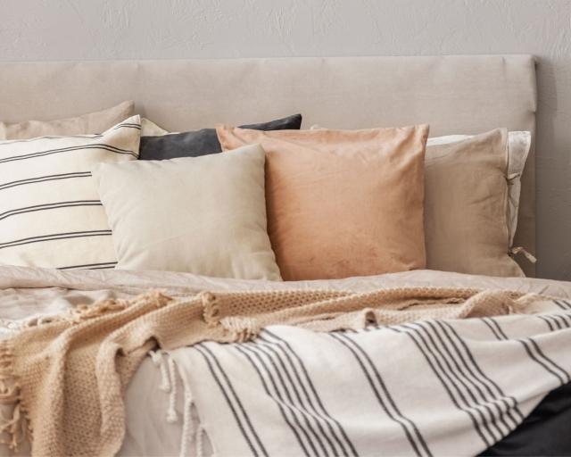 A cozy bed with neutral-colored throw pillows and blankets next to a vase of flowers on a bedside table.