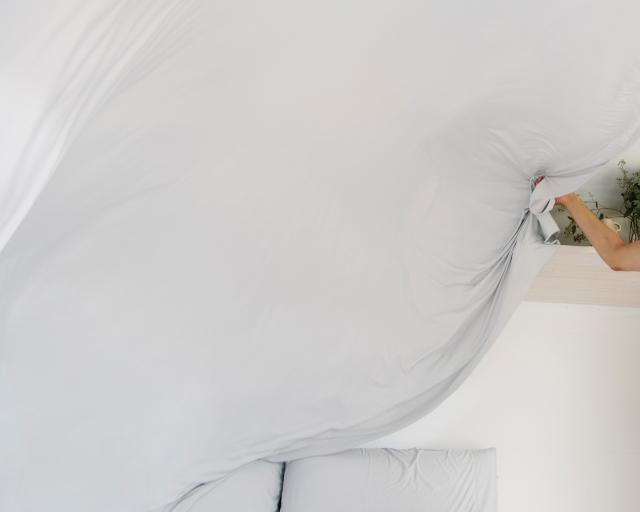 A smiling woman waves a sheet in the air while putting it on a bed.