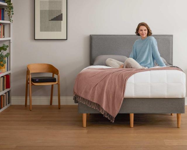 A woman sits upright on top of the covers of a bed on the Purple Bed Frame.