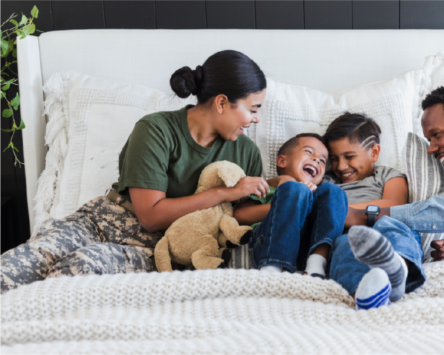 A dark-skinned family, including a female soldier, two young boys, and an adult man, cuddling on a large bed.