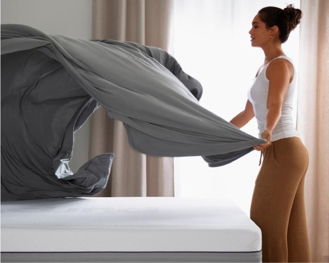 A woman fanning out sheets over a bed.