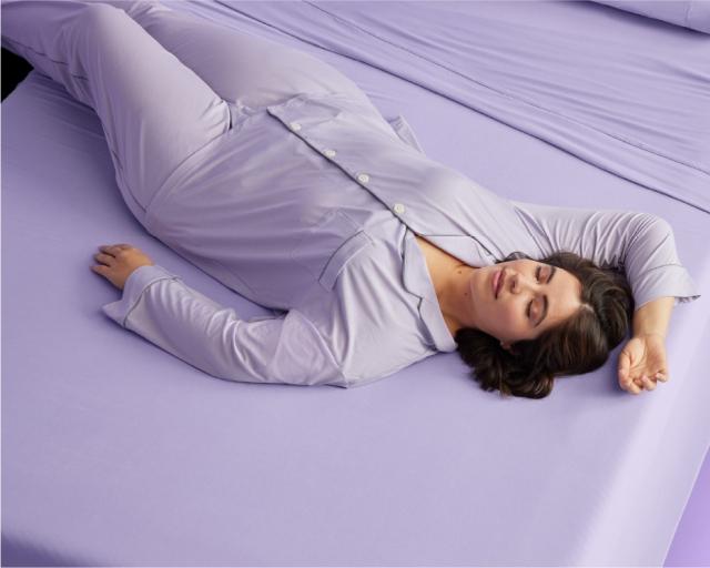 A woman in purple pajamas lying on a bed with purple sheets.