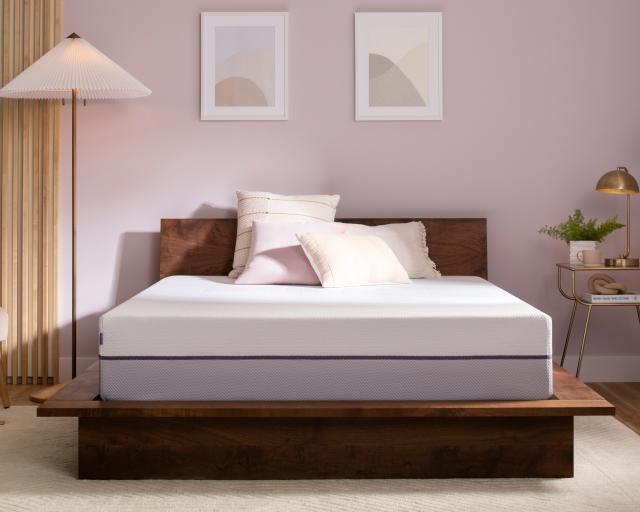Unmade Purple Plus Mattress with throw pillows on a wood bed frame next to a floor lamp, velvet chair, and nightstand.