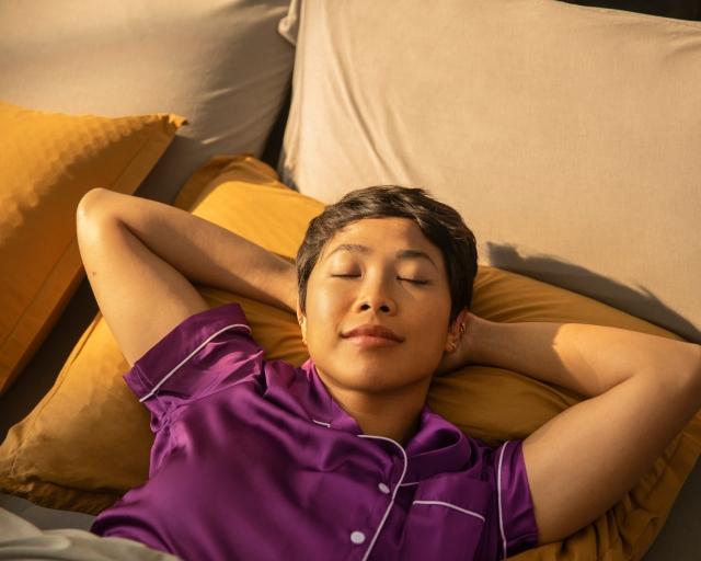 A woman wearing purple satin pajamas rests on a Purple pillow with her arms up behind her head