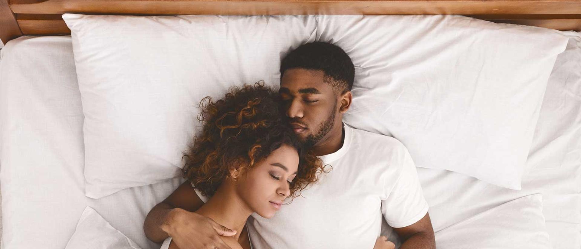 A couple cuddling together in a california king bed with white sheets and white pillows.