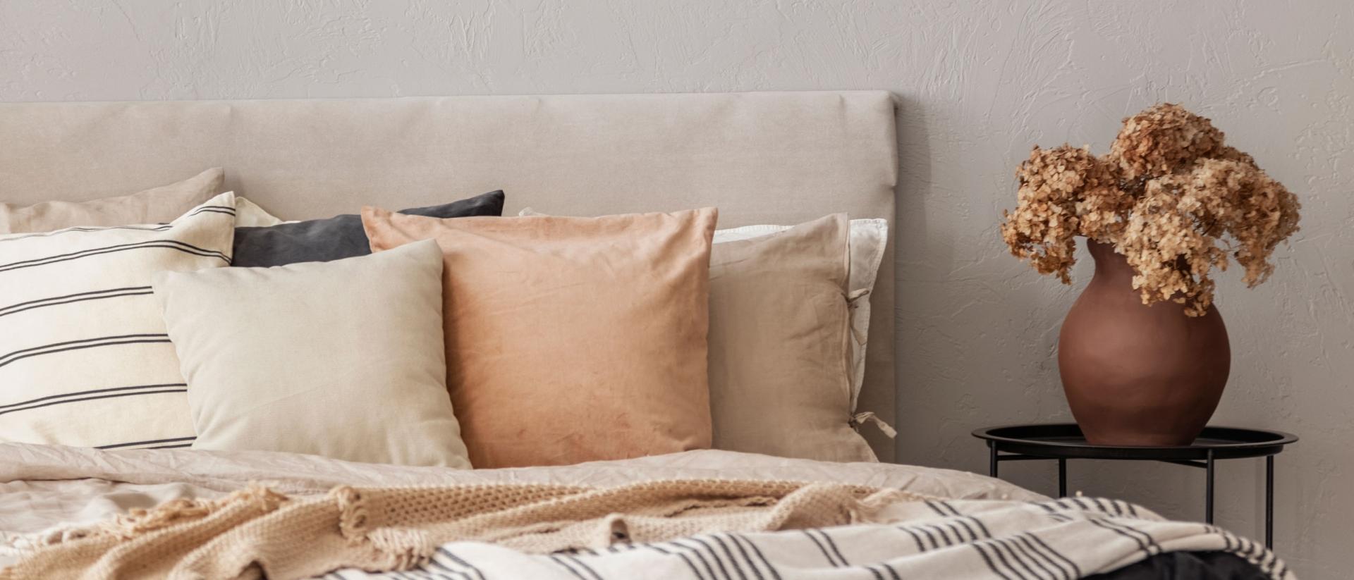 A cozy bed with neutral-colored throw pillows and blankets next to a vase of flowers on a bedside table.