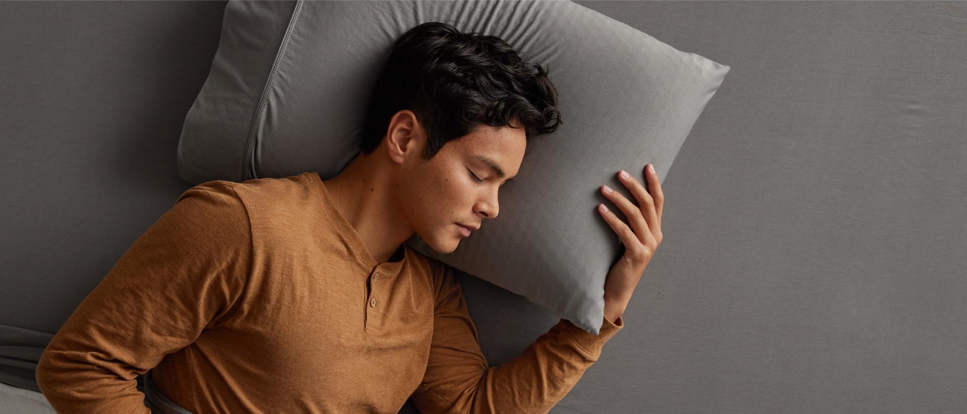 A man falls asleep fast on his side on a bed with gray sheets and white duvet.