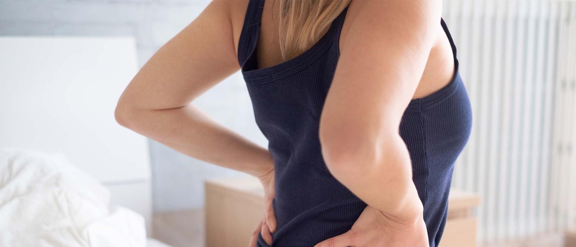 Woman sits on the edge of a bed and holds her back in pain.