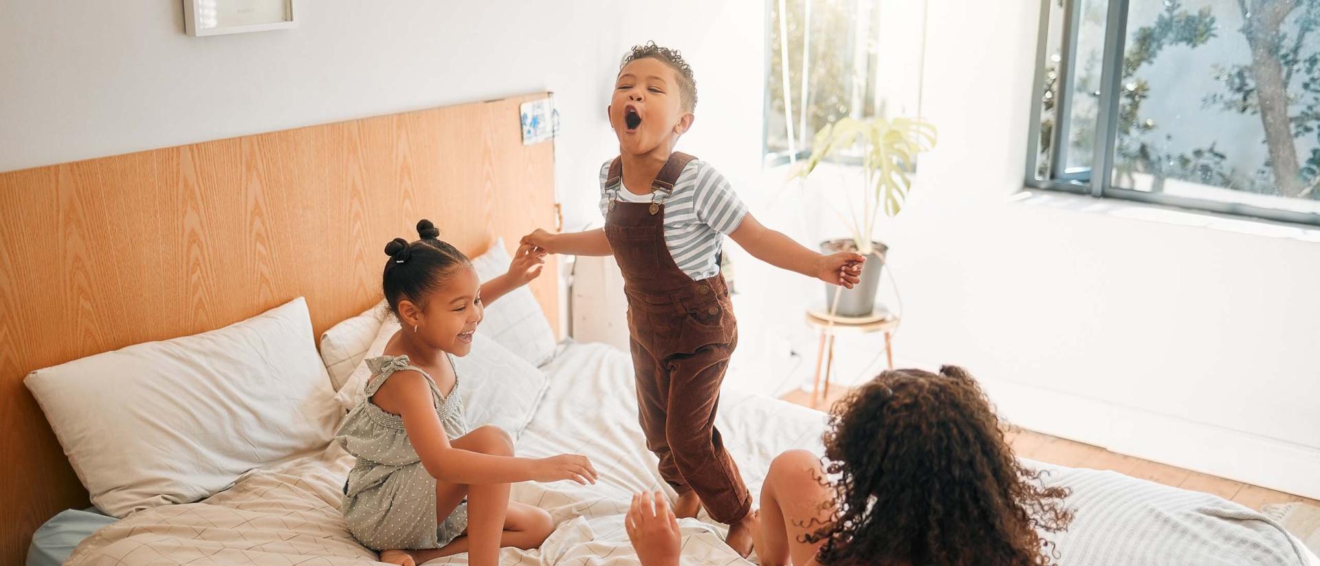 Three young children hold hands while they jump on a bed.