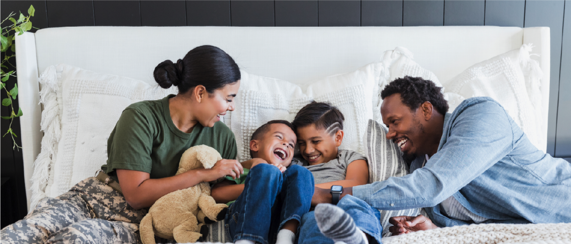 A dark-skinned family, including a female soldier, two young boys, and an adult man, cuddling on a large bed.