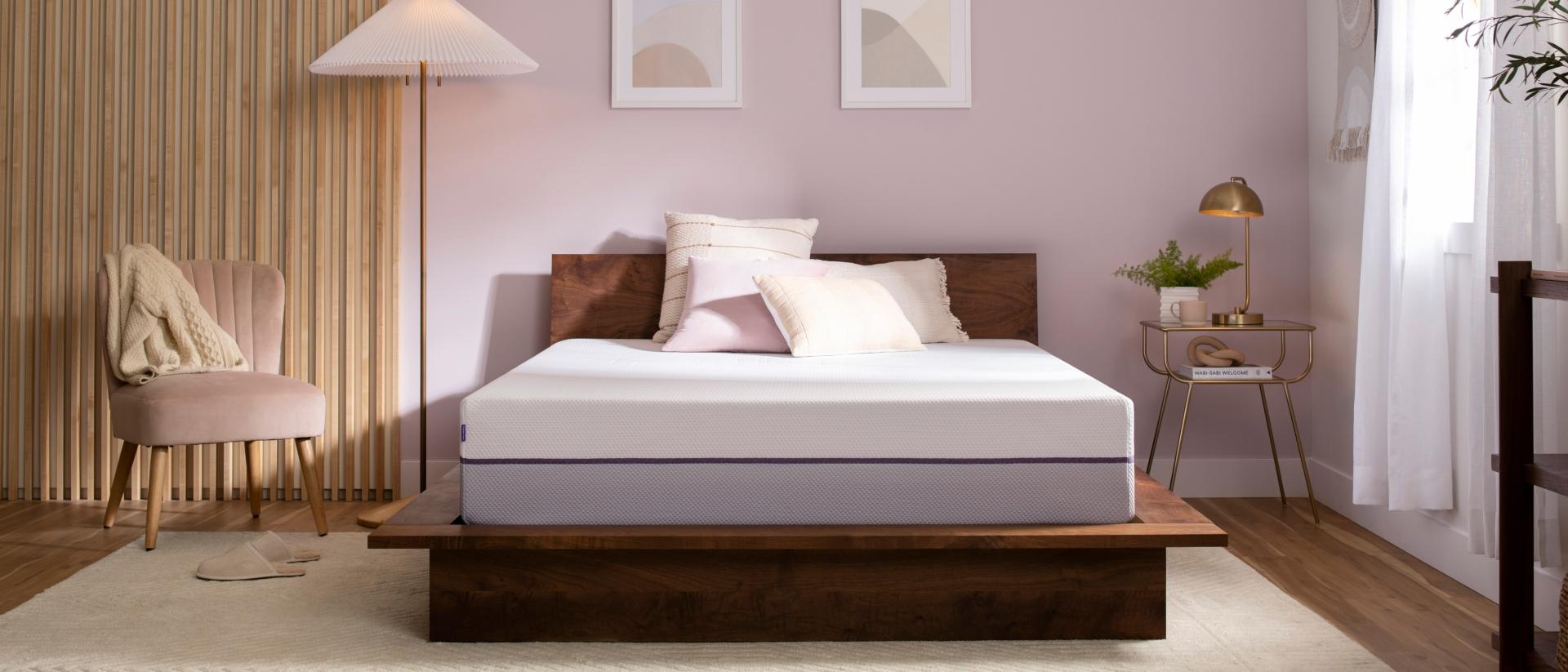 Unmade Purple Plus Mattress with throw pillows on a wood bed frame next to a floor lamp, velvet chair, and nightstand.
