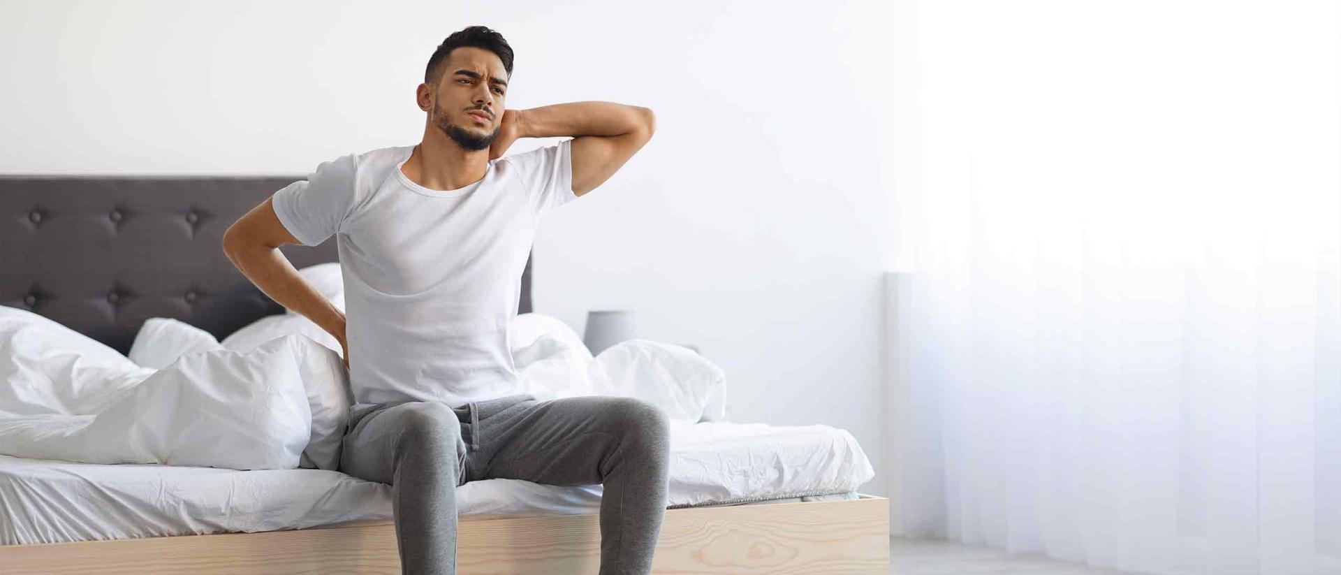 Man sitting on foot of bed in pain after sleeping on a sagging mattress